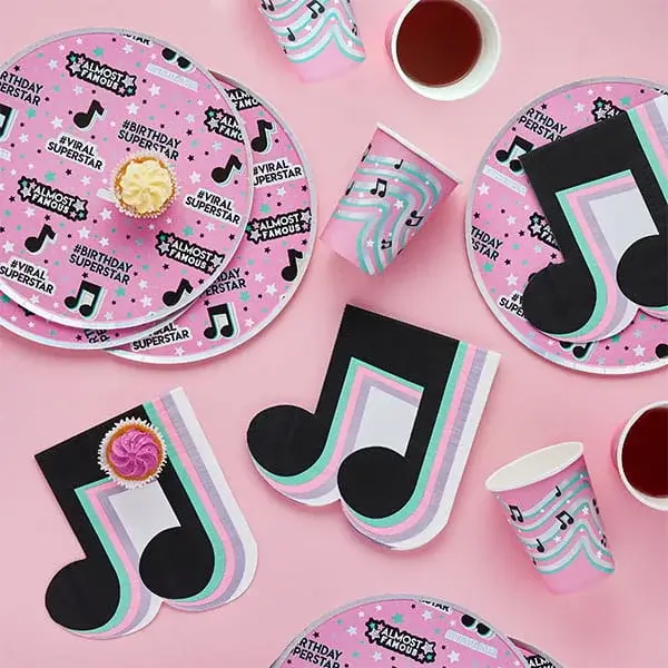 Music-themed party tableware with pink plates, cups, and napkins featuring colorful musical note designs and small cakes